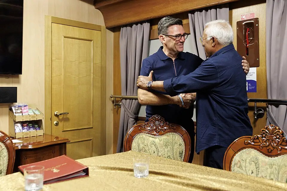 Andy Burnham embraces António Costa, in a wood-pannelled rail carriage behind a covered conference table and upholstered chairs. as they travel by train to Ukraine.